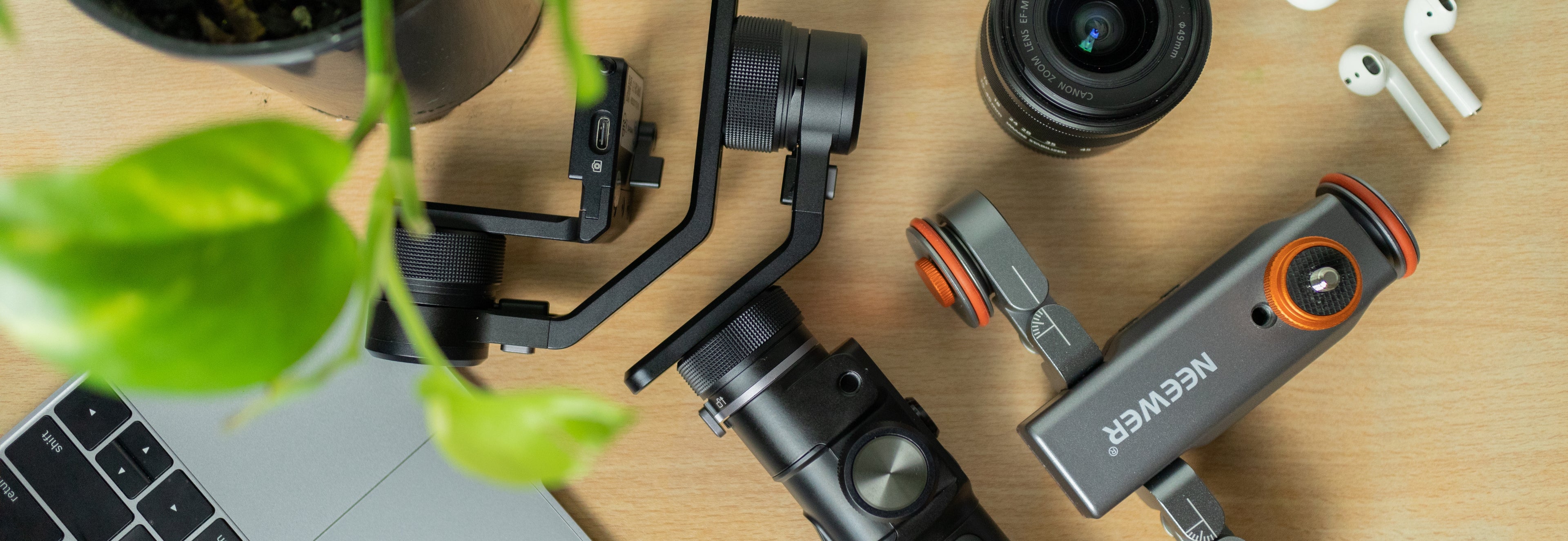 Camera equipment including a laptop, camera stabilizer, and lens on a wooden surface with a plant.