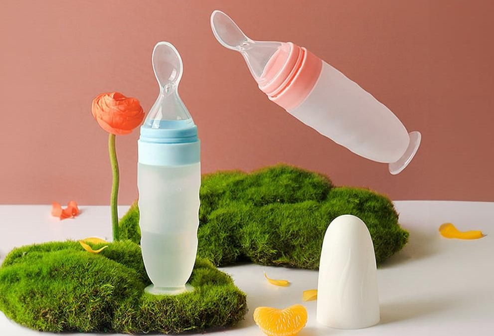 Two baby bottles with spoons on a white surface with green grass and flowers.